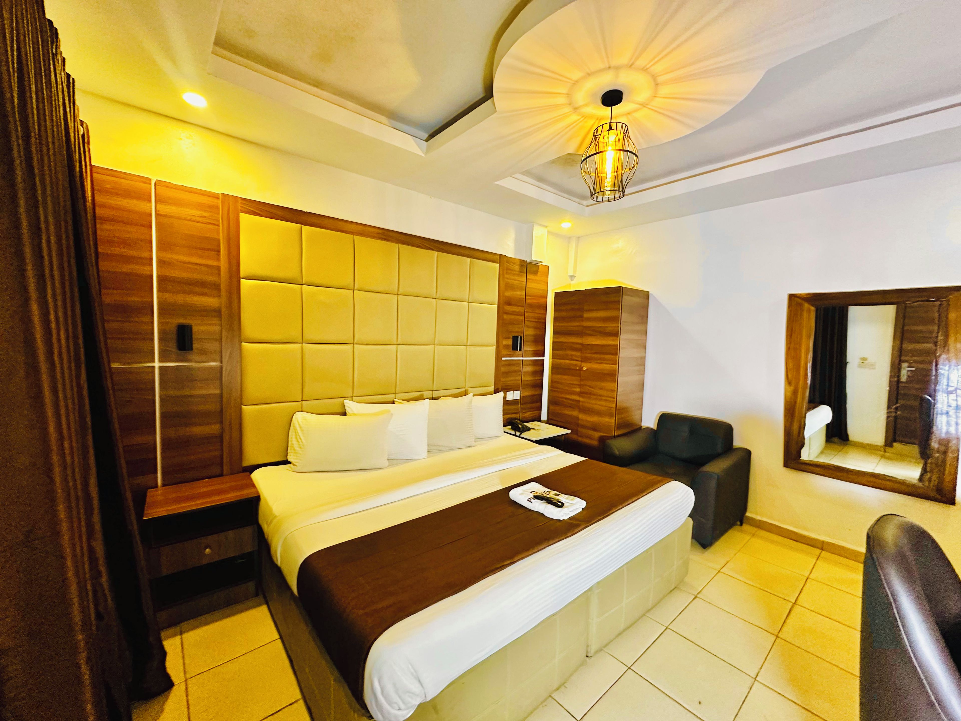 A hotel room with a large bed, a wooden headboard, a bedside table, a chair, a mirror, and a ceiling light fixture.