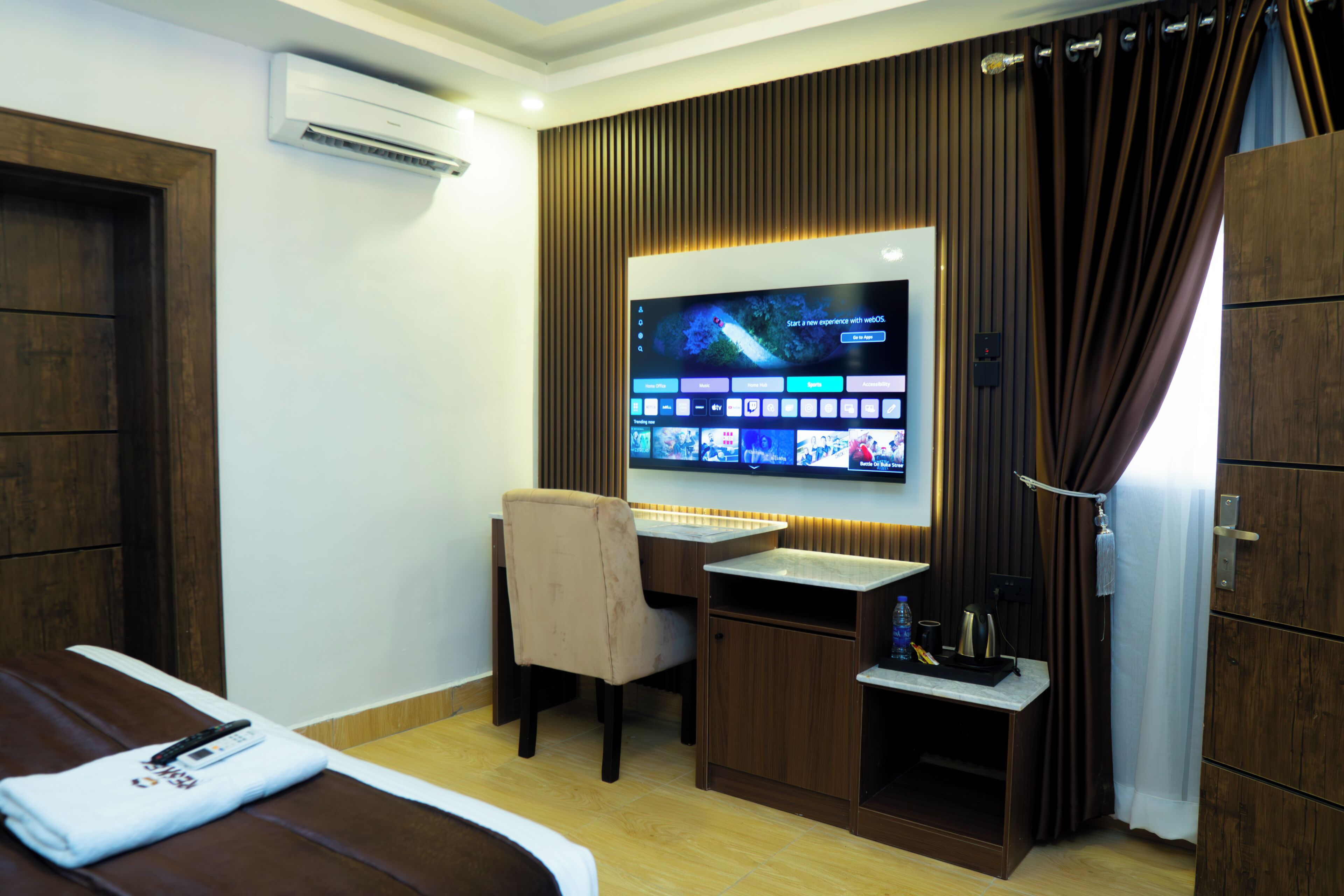 A hotel room with a bed, a desk, a chair, a TV mounted on the wall, and an air conditioning unit.