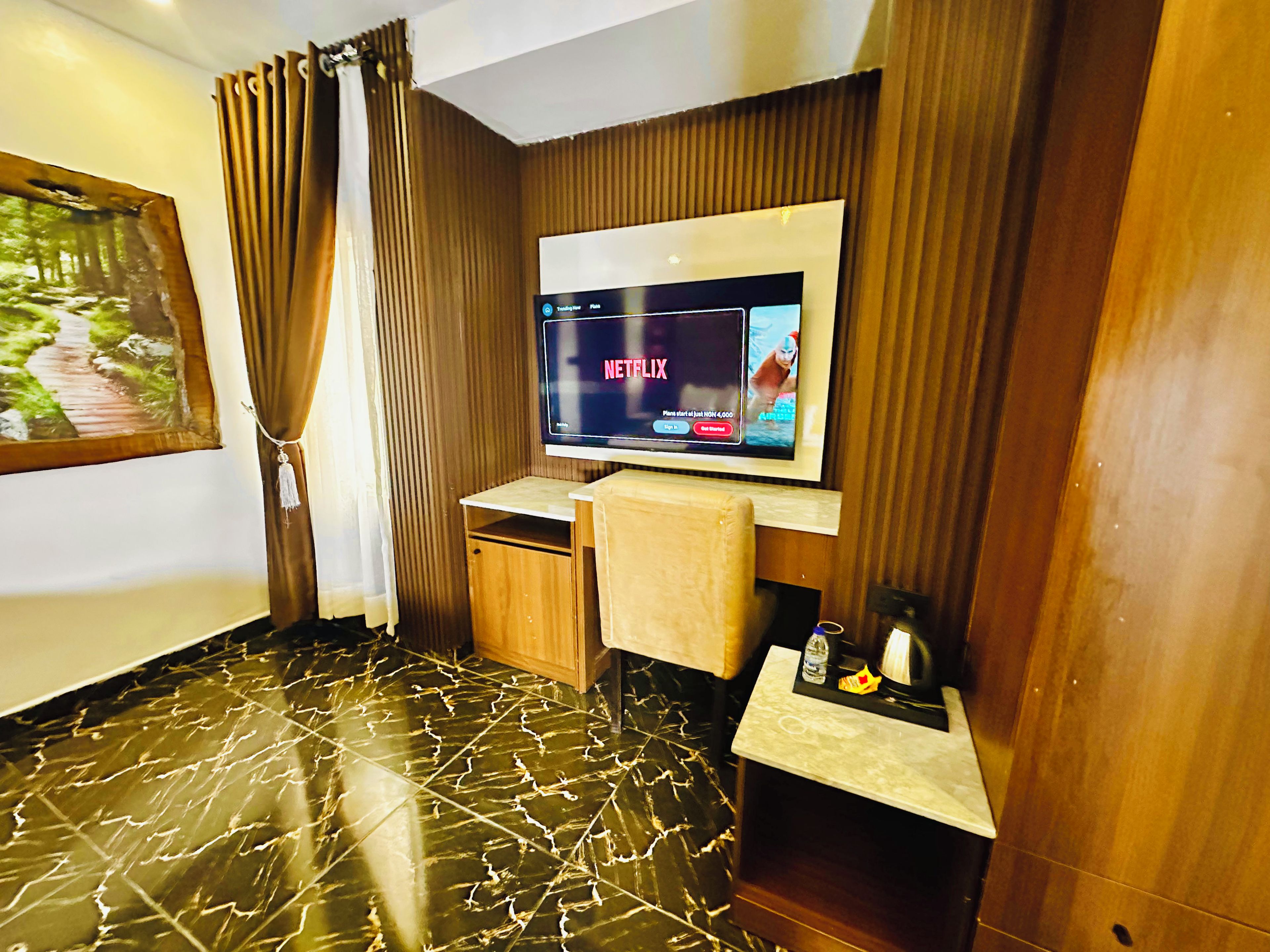 A hotel room with a flat-screen TV mounted on the wall, a wooden cabinet, a small table, and a marble-patterned floor.