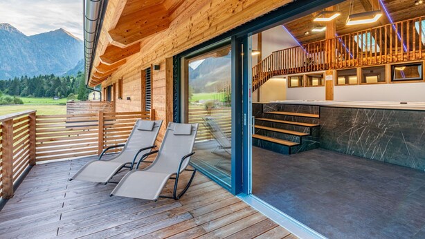Una terraza de madera con dos sillones, una escalera que lleva a un balcón con barandilla y vistas a montañas al fondo.