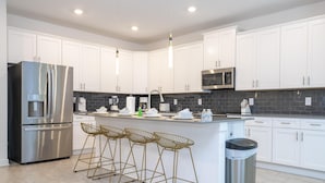 A modern kitchen with white cabinets, stainless steel appliances, a central island with bar stools, and a trash can. - 4342 - Great Family Home near Disney, Pool & Spa - Storey Lake Resort (Kissimmee)