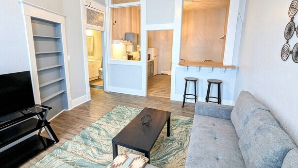 Un salon avec un canapé gris, une table basse noire, un meuble pour la télévision et un tapis à motifs. On aperçoit deux tabourets et un coin cuisine à l’arrière-plan.