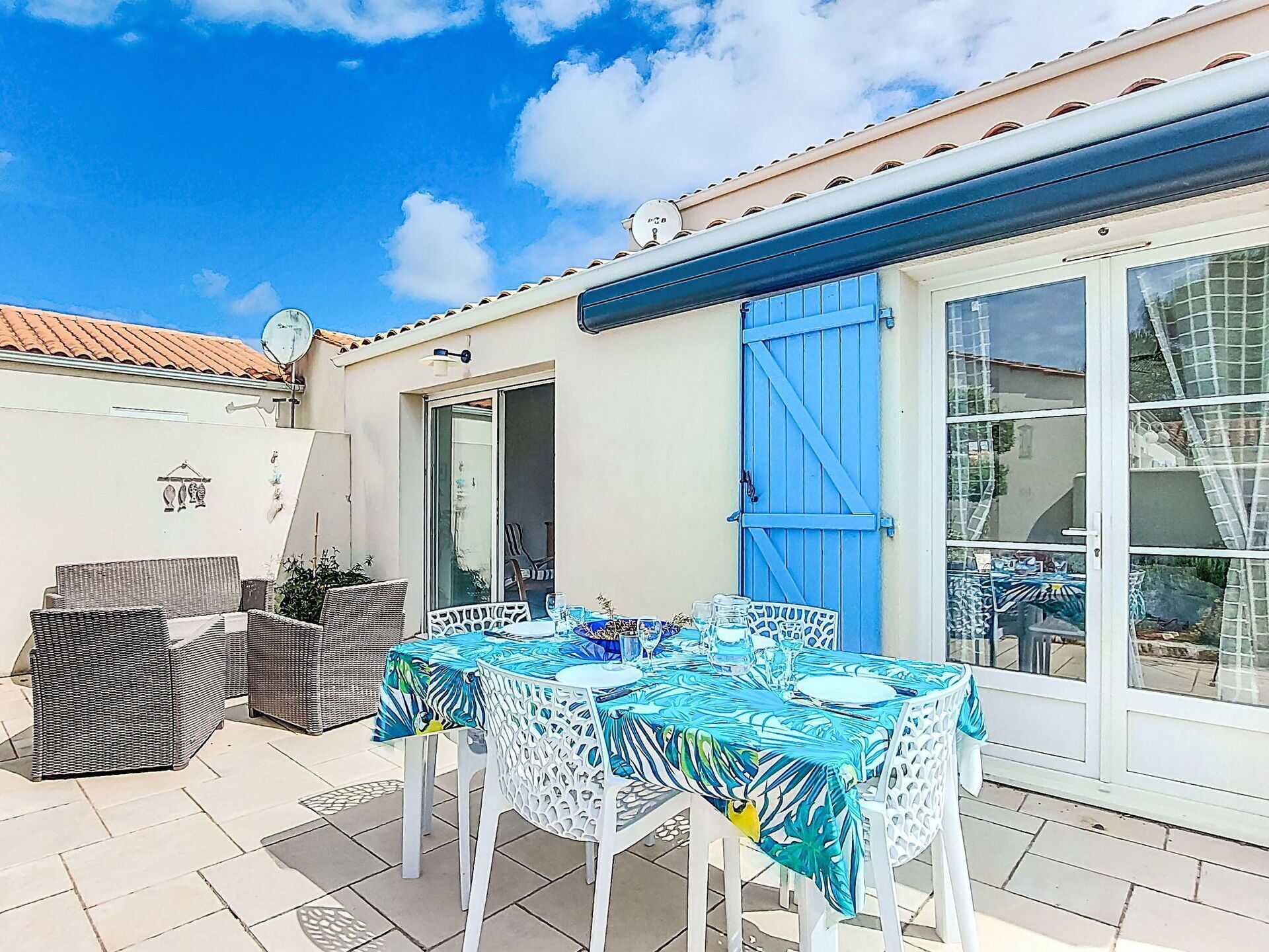 Eine Terrasse im Freien mit Tisch für zwei Personen, weißen Korbmöbeln und einer blauen Tür.