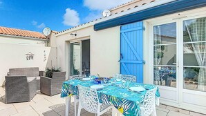 Eine Terrasse im Freien mit Tisch für zwei Personen, weißen Korbmöbeln und einer blauen Tür.