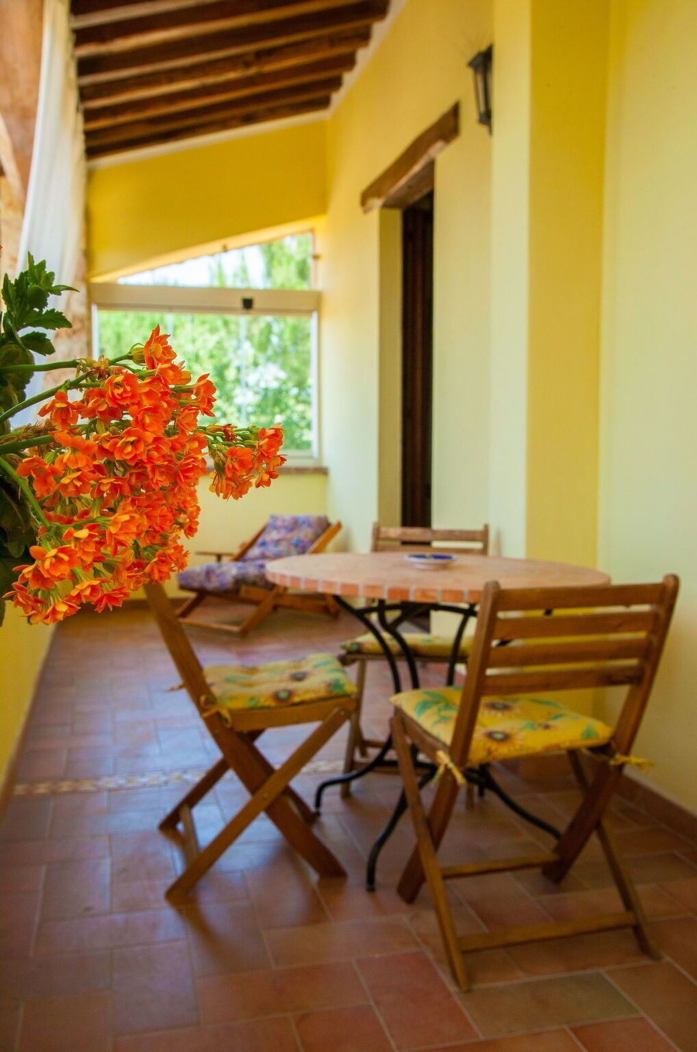 A dining area with a round table and four wooden chairs, a potted plant with orange flowers, and a window with a view of greenery.