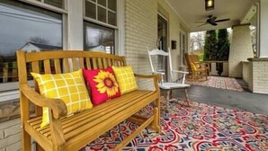 A wooden bench with yellow and red cushions, a white chair, and a patterned rug on a porch with a brick wall and windows. - Central Charleston Craftsman (Charleston)