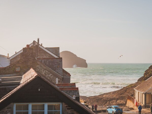 A coastal building with a stone facade and a steep roof, overlooking the sea with waves crashing against a rocky outcrop.