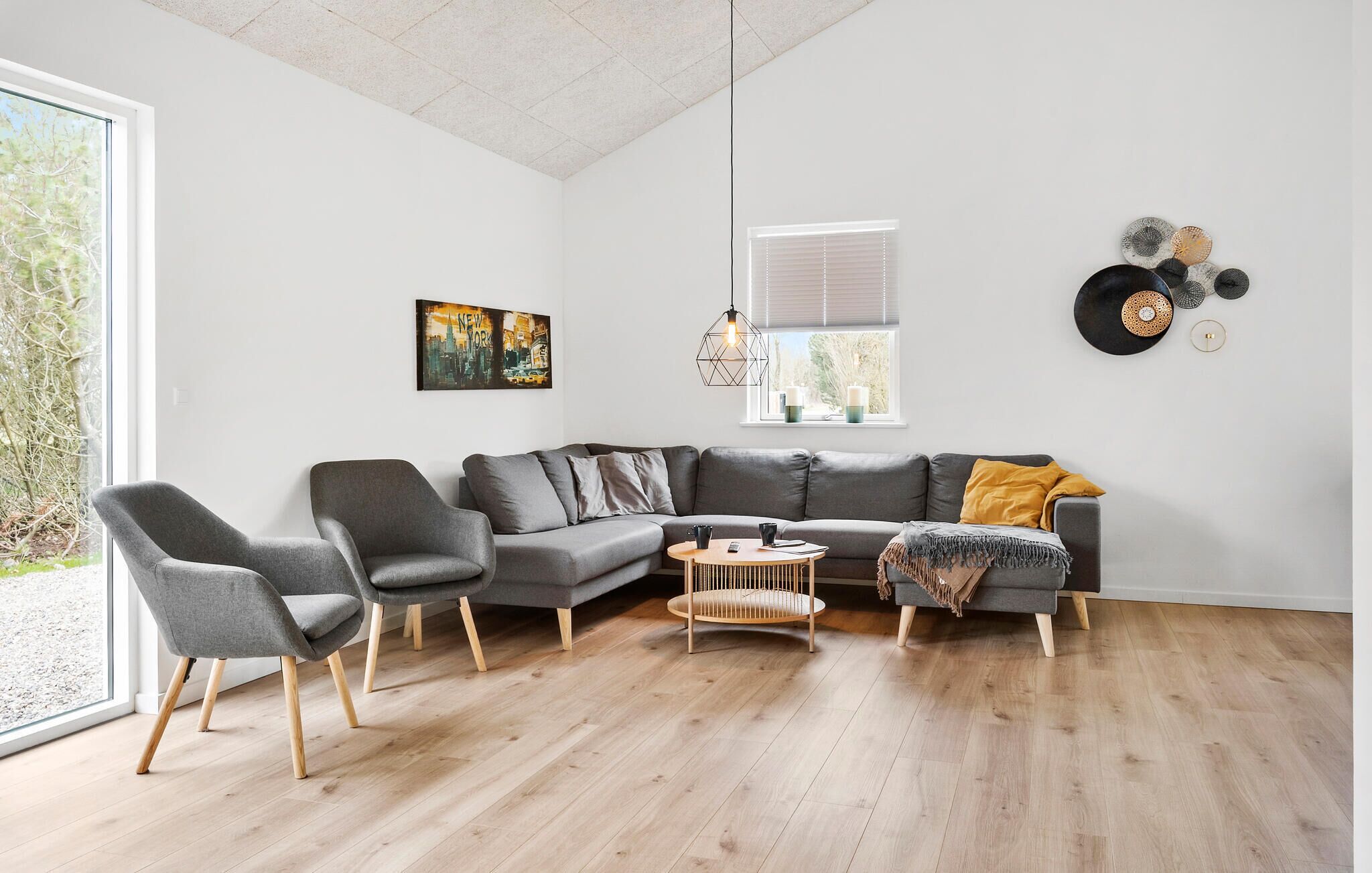 Ein modernes Wohnzimmer mit einem grauen Ecksofa, einem Holztisch und einem grauen Sessel. An der Wand hängt ein Stadtbild und ein Plattenspieler mit Schallplatten steht bereit.
