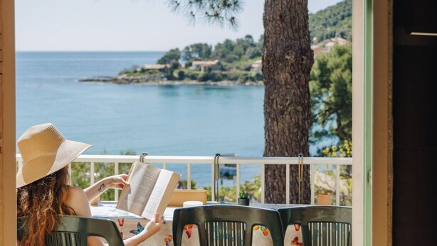 A person reading a book at a table with a view of the sea and a tree. - Lovely apartment for 6 guests with A/C, pool, WIFI, terrace, pets allowed and panoramic view (Tarco)