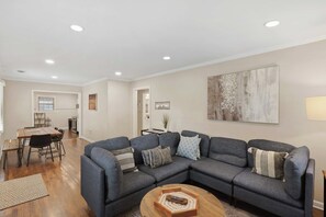 A modern living room with a sectional sofa, a dining area with a table and chairs, and a kitchen in the background.