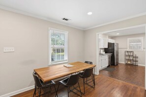 A dining area with a wooden table and chairs, a kitchen with white cabinets and appliances, and a window with blinds.