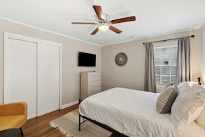 A bedroom with a bed, a ceiling fan, a TV, a dresser, a window with curtains, and a door.