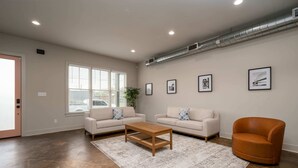 Ein geräumiges Wohnzimmer mit zwei Sofas, einem Couchtisch und einem Sessel. An der Wand befinden sich gerahmte Bilder und ein großes Fenster mit Jalousien.