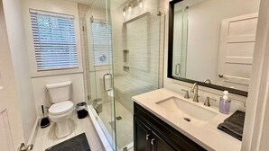 Un baño moderno con cabina de ducha de vidrio, inodoro blanco, lavamanos con espejo y ventana con persianas.