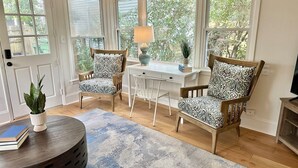 Una habitación con dos sillones estampados, un escritorio blanco, una lámpara, una mesa de centro redonda y vistas a la vegetación a través de ventanas grandes.