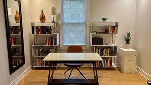 Una habitación con una mesa blanca, dos estanterías, un espejo, una ventana con persianas y un mueble pequeño con una planta.
