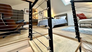 Un dormitorio moderno con cama, una silla y un escritorio. Se observa una escalera con barandilla y un tragaluz arriba.