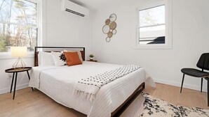 A bedroom with a large bed, a bedside table with a lamp, a chair, and a wall-mounted air conditioner.