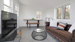 A modern living room with a flat-screen TV, a round glass coffee table, a brown leather sofa, and a wooden bench.