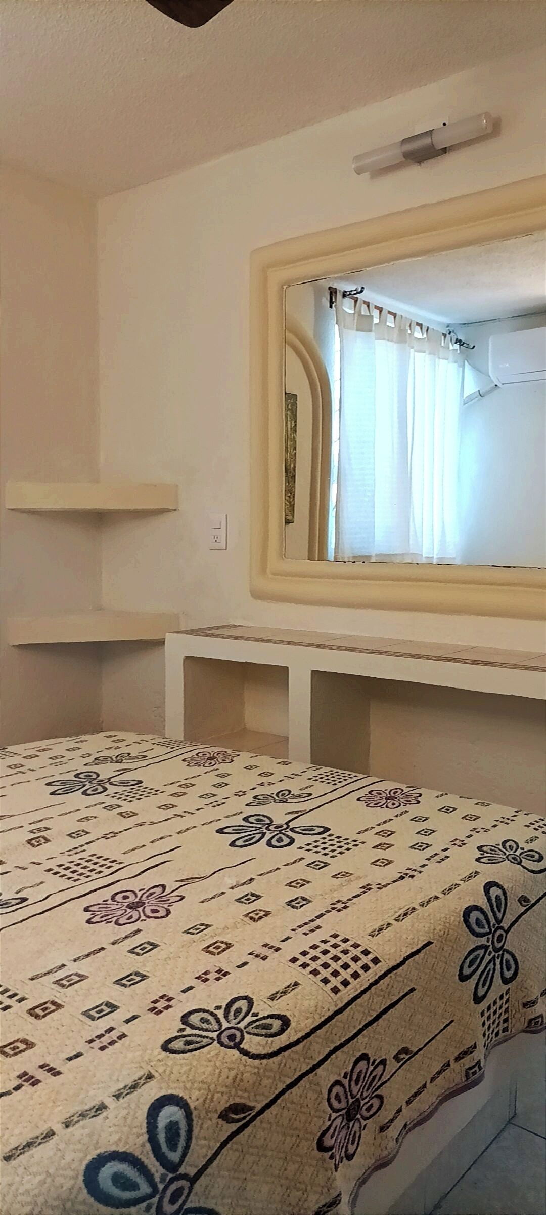 A bedroom with a bed covered in a patterned bedspread, a bedside table, a mirror, and a window with curtains.