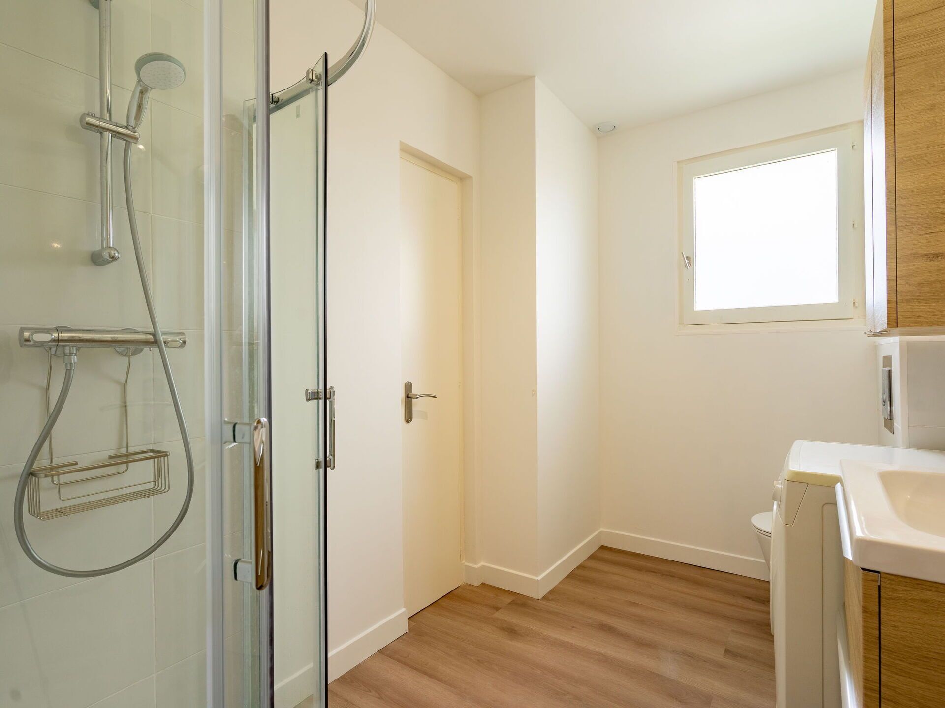 A bathroom with a glass shower enclosure, a white door, a wooden floor, and a white sink.