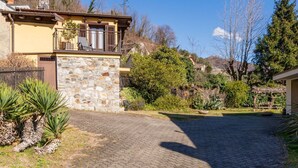 A stone building with a balcony, surrounded by lush greenery and a cobblestone driveway.