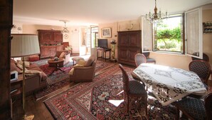 A traditional living room with a dining area, featuring wooden furniture, a chandelier, and a large window with a view of greenery.