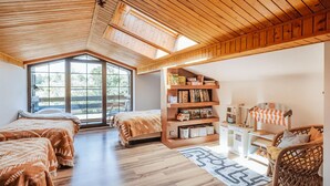 Une chambre mansardée accueillante, avec deux lits, une bibliothèque, une petite table et une fenêtre donnant sur les arbres.