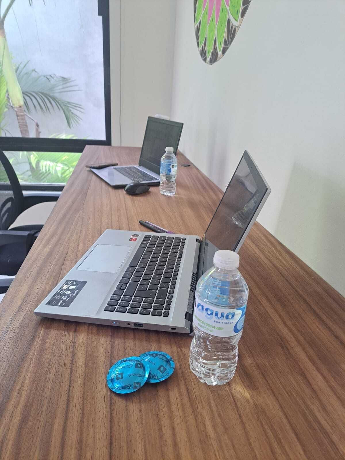 A wooden desk with two laptops, a water bottle, and a pair of blue coasters.
