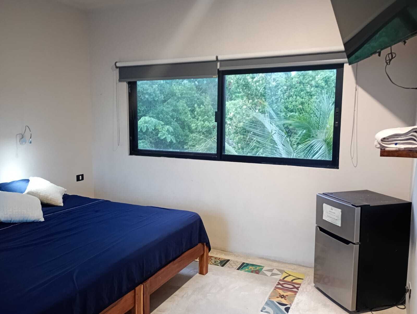 Une chambre avec un lit, une fenêtre donnant sur un espace verdoyant et un réfrigérateur.