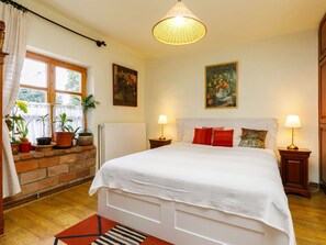 A bedroom with a large bed, two bedside tables with lamps, a radiator, a window with curtains, and a hanging light fixture.
