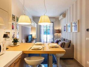 A modern kitchen with a dining table, two pendant lights, a TV, and a sofa in the background.