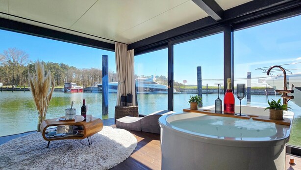 A modern bathroom with a large bathtub, a wooden side table with toiletries, and a view of boats on a river. - Beautiful ship in Sagard with sauna (Sagard)
