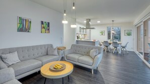 Ein modernes Wohnzimmer mit grauen Sofas, einem runden Holztisch und einem Essbereich, mit einer Küche im Hintergrund.