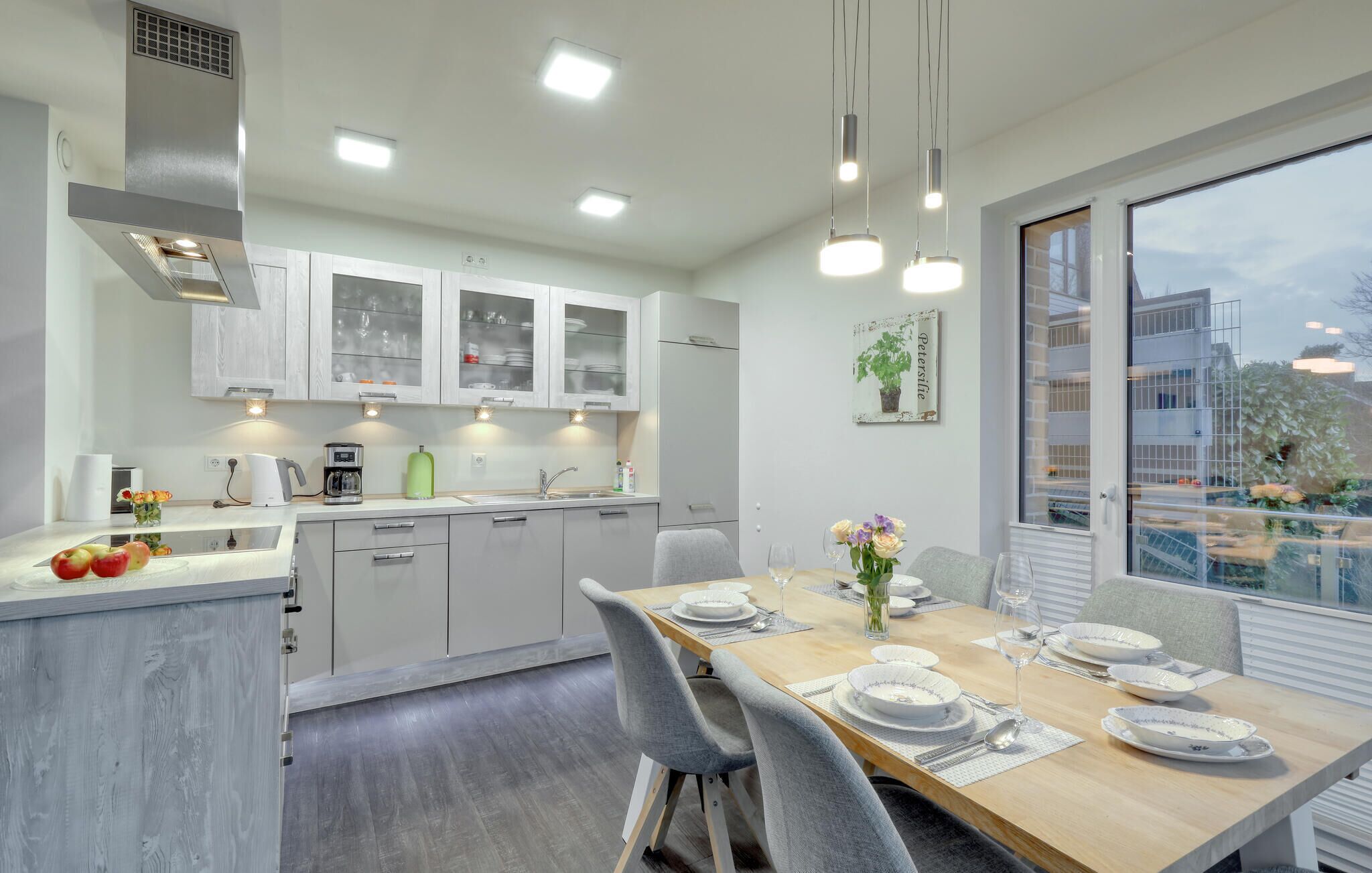 Una cocina moderna con armarios blancos, un comedor con una mesa de madera dispuesta para cuatro personas, y un ventanal que ofrece vistas a edificios y áreas verdes.