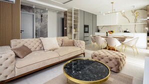 Een moderne woonkamer met een beige bank, een ronde salontafel van zwart marmer, en een eethoek met een witte tafel en gouden stoelen.