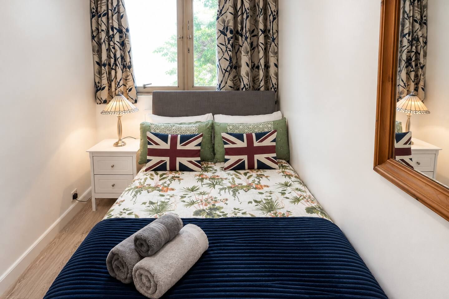A bedroom with a bed, two pillows with Union Jack designs, a bedside table with a lamp, and a mirror on the wall.
