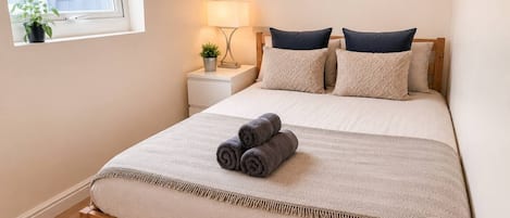 A bedroom with a bed, pillows, a bedside lamp, a nightstand, a window with a plant, and a painting on the wall.