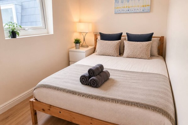 Une chambre à coucher avec un lit, des oreillers, une lampe de chevet, une table de chevet, une fenêtre avec une plante et un tableau au mur.