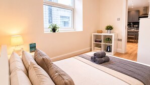A bedroom with a bed, bedside table, lamp, and a window with a view of buildings. - 3bdr Flat Near Aldgate, Brick lane & tower hill (London)