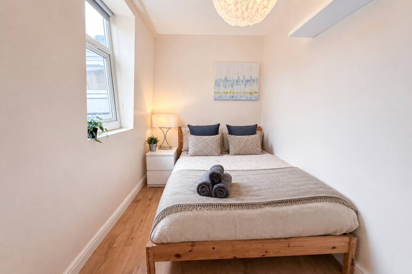 A bedroom with a bed, a nightstand, a lamp, a window, and a painting on the wall.