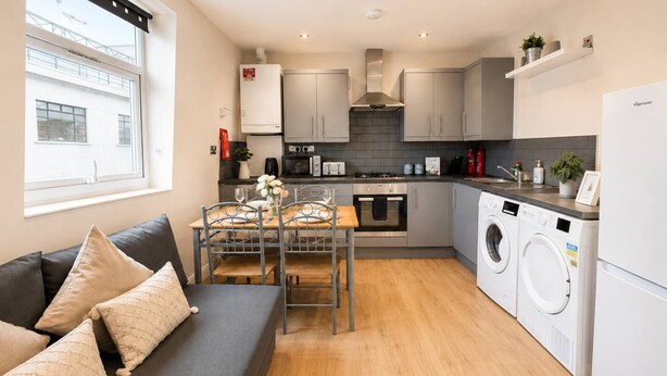 A compact kitchen with a dining area, a washing machine, and a sofa with cushions. - 3bdr Flat Near Aldgate, Brick lane & tower hill (London)