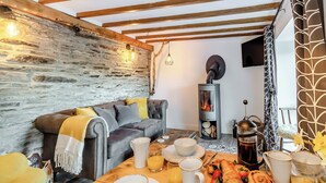 Une pièce chaleureuse avec un mur en pierre, un canapé, une table où est servi le petit-déjeuner et un poêle à bois.