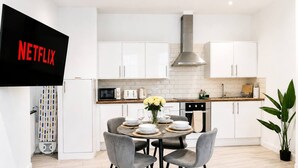 Cozinha moderna com armários brancos, uma TV preta exibindo 'NETFLIX', uma mesa de jantar com cadeiras e um vaso de planta.