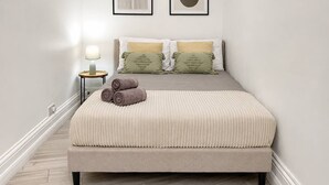 Cama bem arrumada com cobertura bege, duas almofadas e toalhas enroladas sobre um piso de madeira.