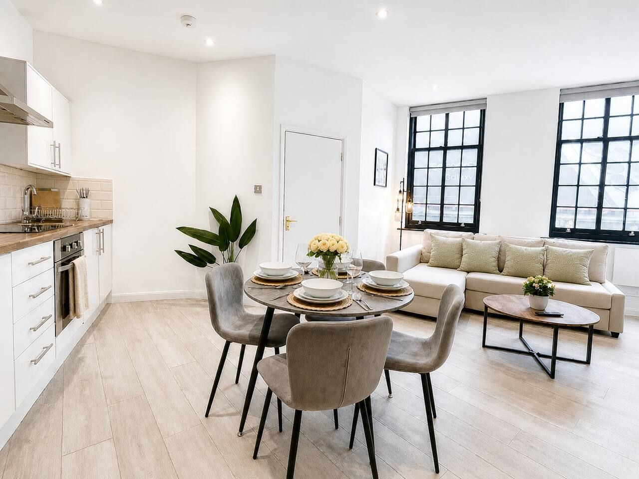 Een moderne keuken met witte kasten, een eethoek met een ronde tafel en stoelen, en een woonkamer met een bank en grote ramen.