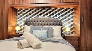 Een net opgemaakt bed met een gestoffeerd hoofdbord, gestreepte kussens en een opgerolde handdoek.