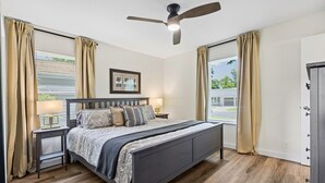 Ein Schlafzimmer mit Bett, Nachttischen, Deckenventilator und großen Fenstern mit Vorhängen.