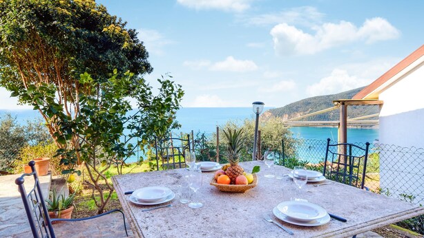 Une table à manger installée à l’extérieur, avec de l’assiette, des couverts et un panier de fruits, offrant une vue sur la côte et un pont en arrière-plan.