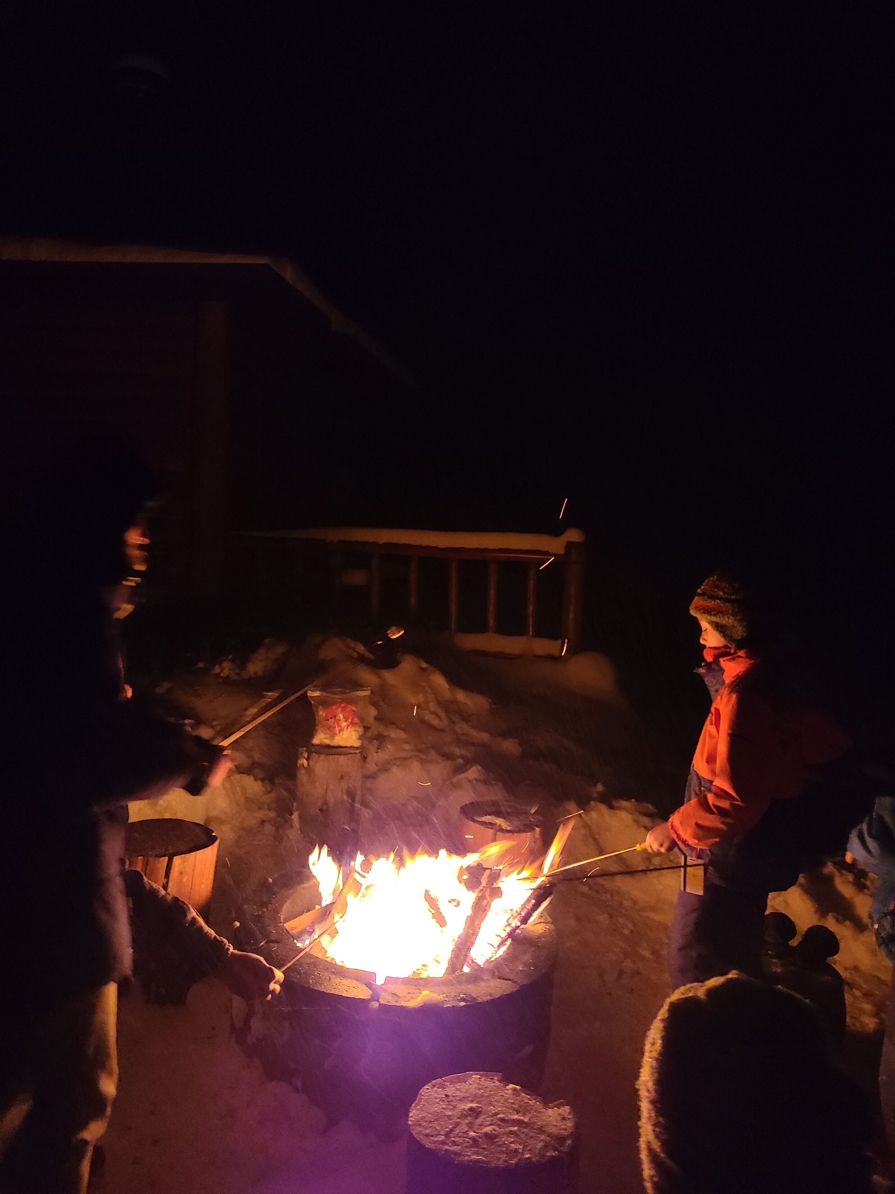 A person cooking outdoors at night with a fire, surrounded by snow.